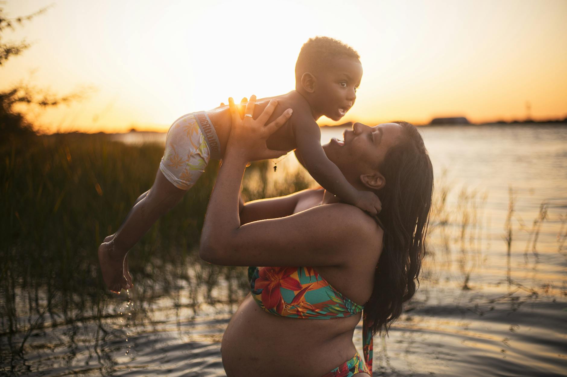 Ciężarna mama trzyma małe dziecko nad wodą o zachodzie słońca